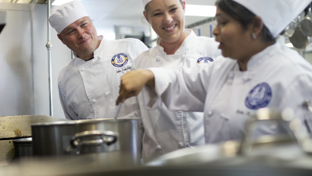 People wearing chef's jacket and hats, one individually is stirring the pot and speaking while the other two watch and smile