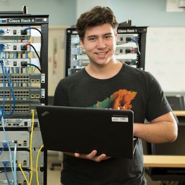 male student holding laptop next to computer servers.
