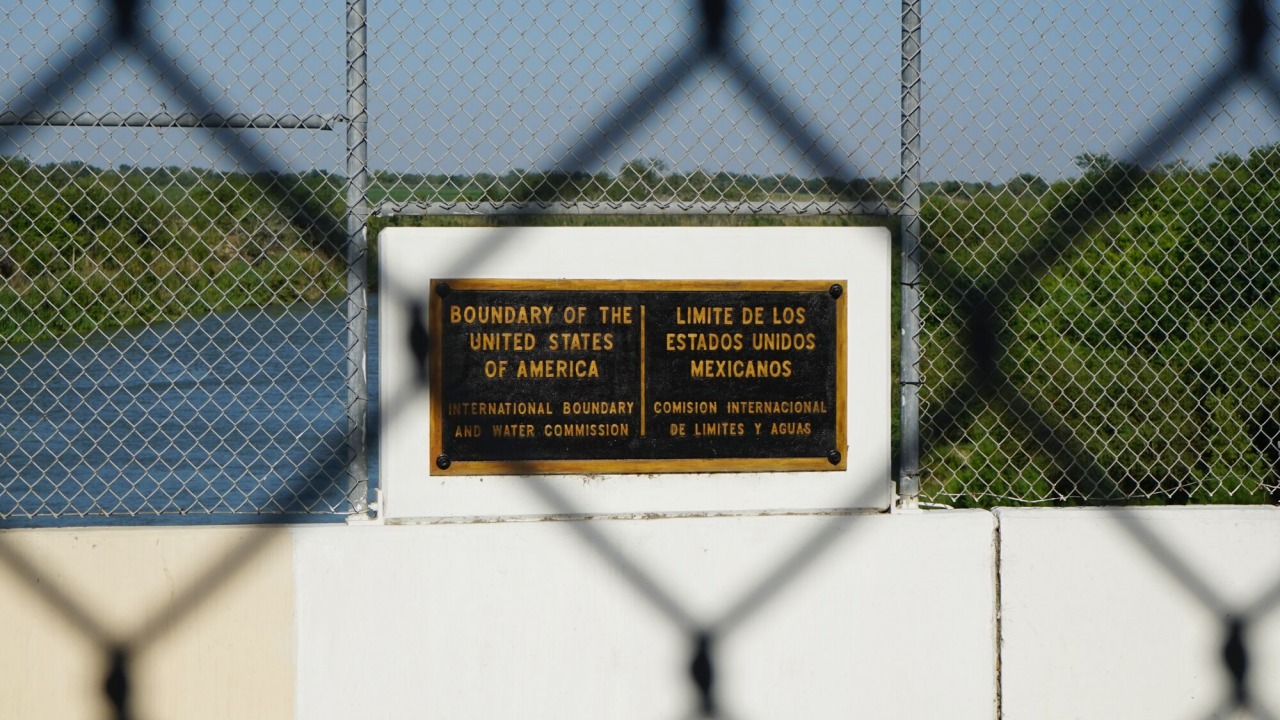 A chainlink fence with a sign that reads "boundary of the United States of America" seen through another chainlink fence