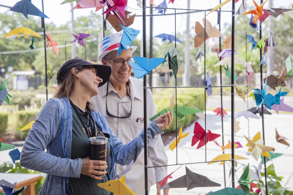 Two people wearing hats looking at various folded paper animals hanging from wire frame