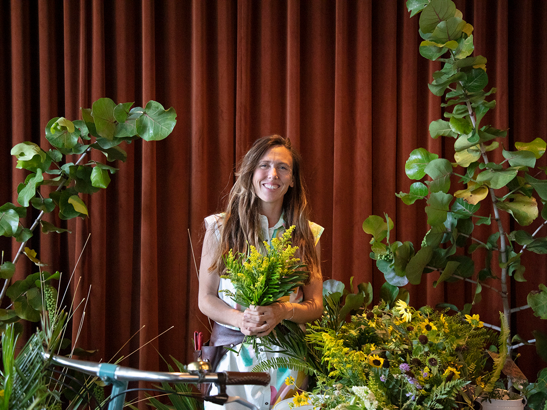 A woman holding a bouquet stands amidst green plants in front of red curtains.