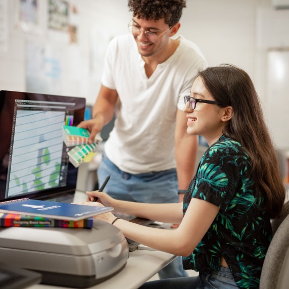 A male design student comparing swatches with his female classmate while they both look at her monitor