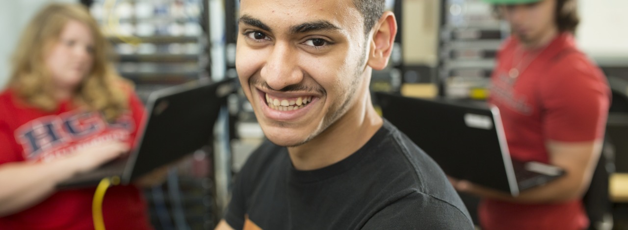 Male student smiling in front of two students using laptops standing in front of server racks