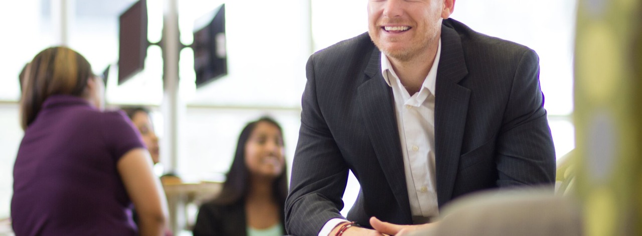 Smiling white adult male wearing a business suit sitting in a lounge leaning forward with students in the background