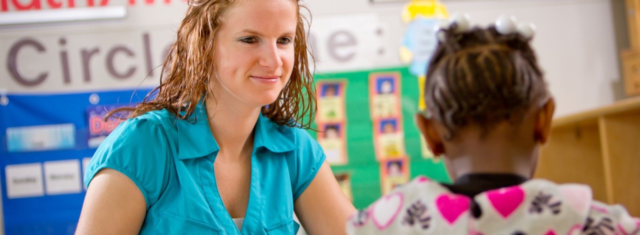 White female adult sitting and smiling at a table with a young African-American girl in a classroom