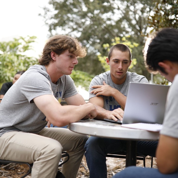 Three male students sitting at a table outside working on a laptop.